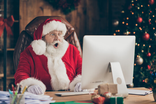 Photo of amazed santa claus sit table desk work computer impressed many wish gift list letters in house idnoors with evergreen christmas x-mas tree ornament decoration wear cap headwear
