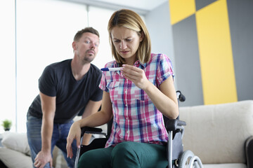 Woman in wheelchair looks closely at pregnancy test from behind man peeks at. Expectation and...