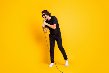 Full length photo of young man hold mic sing open mouth wear black t-shirt pants white sneakers spectacles isolated yellow color background
