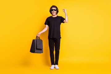 Full length photo of young man stand raise arm fist hold bags wear black t-shirt pants white sneakers spectacles isolated yellow color background