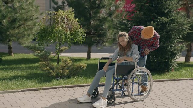 Young man tie shoelaces to his disabled invalid girlfriend on wheelchair. Couple in love walking outside. Touching moment of help and support. Concept of disability assistance. 6k downscale 10 bit
