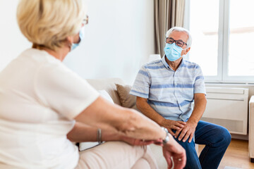 Senior friends having conversation on the sofa at home during Covid 19 pandemic. Elderly wearing medical face mask