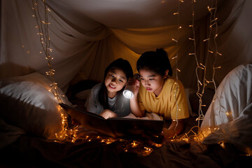 Obraz na płótnie Canvas Family concept. elder sister and sister reading book with flashlight together before bedtime. Sister read story book together in bed sheet tent. focus selective little sister. With film grain effect