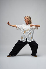 Mature blonde woman practicing Chi kung and Tai Chi on a white background. She wears a traditional white chinese Tai Chi jacket, black trousers and black shoes with ying yang symbol