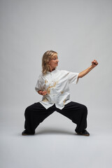 Mature blonde woman practicing Chi kung and Tai Chi on a white background. She wears a traditional white chinese Tai Chi jacket, black trousers and black shoes with ying yang symbol