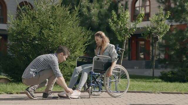 Young man tie shoelaces to his disabled invalid girlfriend on wheelchair. Touching moment of help and support. Couple in love walking outside. Concept of disability assistance. 6k downscale 10 bit
