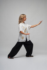 Mature blonde woman practicing Chi kung and Tai Chi on a white background. She wears a traditional white chinese Tai Chi jacket, black trousers and black shoes with ying yang symbol