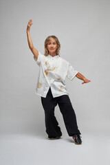 Mature blonde woman practicing Chi kung and Tai Chi on a white background. She wears a traditional white chinese Tai Chi jacket, black trousers and black shoes with ying yang symbol