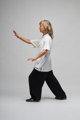 Mature blonde woman practicing Chi kung and Tai Chi on a white background. She wears a traditional white chinese Tai Chi jacket, black trousers and black shoes with ying yang symbol