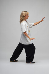 Mature blonde woman practicing Chi kung and Tai Chi on a white background. She wears a traditional white chinese Tai Chi jacket, black trousers and black shoes with ying yang symbol