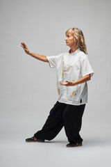 Mature blonde woman practicing Chi kung and Tai Chi on a white background. She wears a traditional white chinese Tai Chi jacket, black trousers and black shoes with ying yang symbol