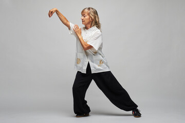 Mature blonde woman practicing Chi kung and Tai Chi on a white background. She wears a traditional white chinese Tai Chi jacket, black trousers and black shoes with ying yang symbol