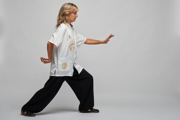 Mature blonde woman practicing Chi kung and Tai Chi on a white background. She wears a traditional white chinese Tai Chi jacket, black trousers and black shoes with ying yang symbol