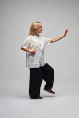 Mature blonde woman practicing Chi kung and Tai Chi on a white background. She wears a traditional white chinese Tai Chi jacket, black trousers and black shoes with ying yang symbol