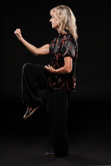 Mature blonde woman practicing Tai Chin Chen style in a black background wearing a traticional chinese black jacket with red decoration, black trousers and traditional shoes with ying yang symbol