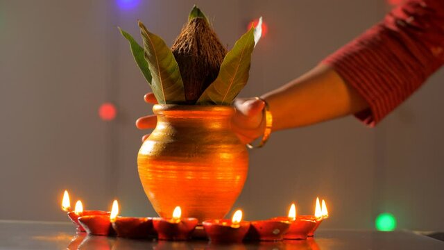 Female's beautiful hand keeping a copper puja Kalash in between some oil lamps. Close-up shot of lighted Diyas and Kalash with coconut and mango leaves in a puja room for Diwali/Hindu prayers