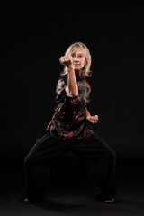 Mature blonde woman practicing Tai Chin Chen style in a black background wearing a traticional chinese black jacket with red decoration, black trousers and traditional shoes with ying yang symbol