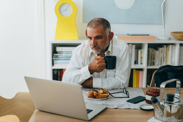 mid aged man having breakfast at home drinking coffee and checking mail on computer