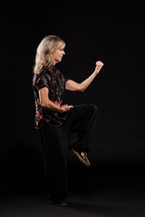 Mature blonde woman practicing Tai Chin Chen style in a black background wearing a traticional chinese black jacket with red decoration, black trousers and traditional shoes with ying yang symbol