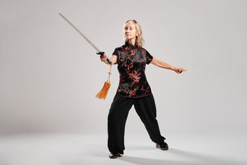 Mature blonde woman practicing Tai Chin with sword in a white background wearing a traditional chinese black jacket with red chinese decoration, black trousers and chinese shoes with ying yang symbol