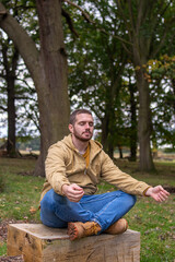 Photo of a young and attractive man sitting on a tree trunk practising meditation in the middle of nature. Self isolation in a forest. Relax