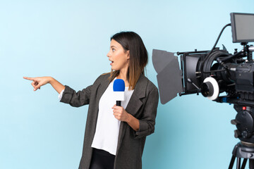 Reporter woman holding a microphone and reporting news over isolated blue background pointing...