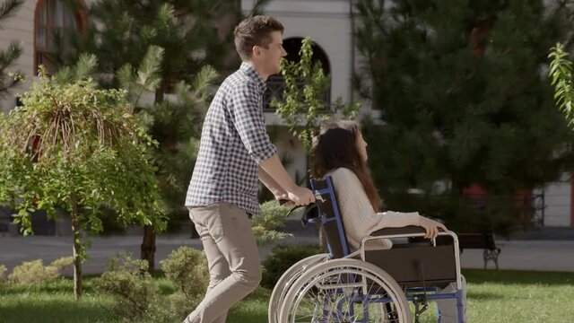Young man riding his disabled invalid girlfriend on wheelchair talking and laughing. Asian handicapped girl with paralysis living full life. Couple in love. 6k downscale 10 bit color.