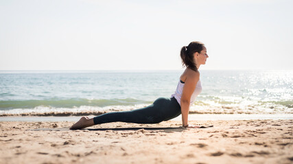 Fototapeta na wymiar Young playful woman doing sport exercises on the beach at sunset along the the Atlantic Ocean coastline. Attractive portuguese woman doing Yoga poses on the beach. Relaxing and meditation lifestyle