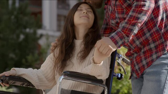 Portrait of disabled happy young woman n wheelchair and her husband kissing her. Asian handicapped girl with paralysis living full life. Couple in love. 6k downscale 10 bit color.