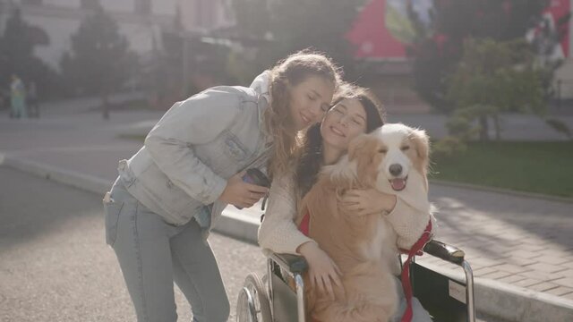 Pretty disabled invalid girl sitting on wheelchair with a guide dog and hugging her friend while riding on the street. Two happy girlfriends spending time outdoors on lovely sunny day.