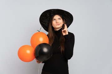 Young witch holding black and orange air balloons with fingers crossing and wishing the best