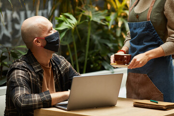 Mature man in protective mask working at the table with laptop while waiter serving the coffee at the restaurant