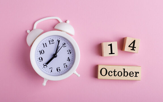 October 14 written on wooden cubes near a white alarm clock on a pink background