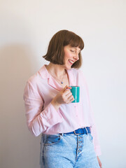 Young woman wearing jeans and pink shirt drinking coffee and smiling over white wall background.