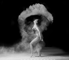beautiful caucasian woman in a black bodysuit with a sports figure is dancing in a white cloud of flour