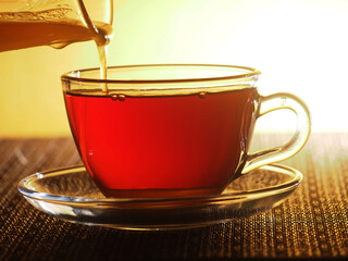 A glass Cup of tea is filled with honey from a glass container.