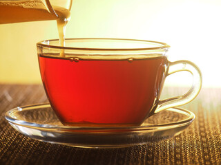 A glass Cup of tea is filled with honey from a glass container.