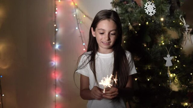 A little girl stands near a Christmas tree holding burning sparklers in her hands