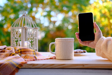 on the table in the autumn garden tea, coffee in a mug, a candle burns, a woman holds a smartphone with a blank black screen, the concept of an outdoor tea party, the use of modern gadgets