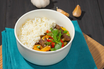 Roast beef with sweet pepper and white rice. White plate, garlic in the background, dark background.
