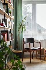 Stylish composition of home office interior with design retro chair, home library, plant, window, books, decoration and elegant personal accessories in home decor.