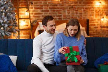 Handsome man presenting a gift to his beautiful wife and smiling. Beautiful young couple at home enjoying spending time together. Winter holidays, Christmas celebrations, New Year concept.