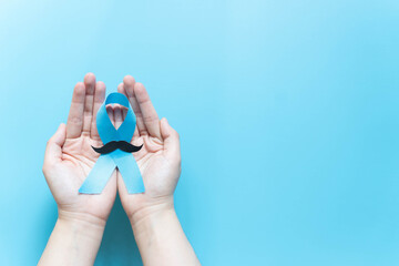 Men's health and Prostate cancer awareness campaign in November. Man hands holding light blue ribbon awareness w/ mustache on blue background. Symbol for support men who living w/ cancer. Copy space.