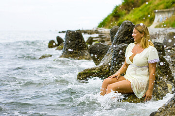A young blonde woman in a short yellow dress sits on large stones, knee-deep in water on the seashore.