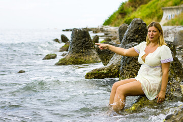 A young blonde woman in a short yellow dress sits on large stones, knee-deep in water on the seashore.