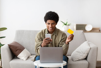 Cool African American teenager with laptop using mobile phone and credit card for online shopping at home