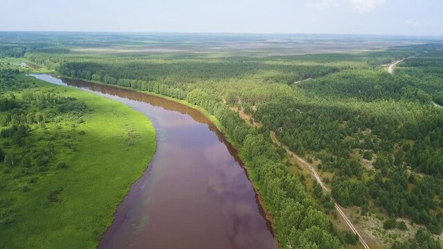 Aerial view of the river and green forest under blue sky in the summer. Clip. Summer landscape with forest, green fields and wide river on a sunny day.