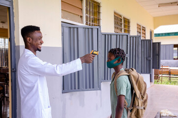 a young handsome african class teacher holding as thermometer to scan the temperature of his student before entering the classroom