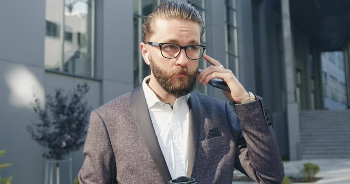Handsome successful serious adult bearded office manager in glasses checking his airpods before starting to talk on smartphone near modern office building