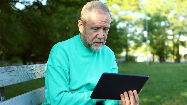 Adult senior man using tablet for online communicating speaking looking at screen resting outside enjoying autumn spring nature. Sunny background. Internet connect. Free communicating with family.